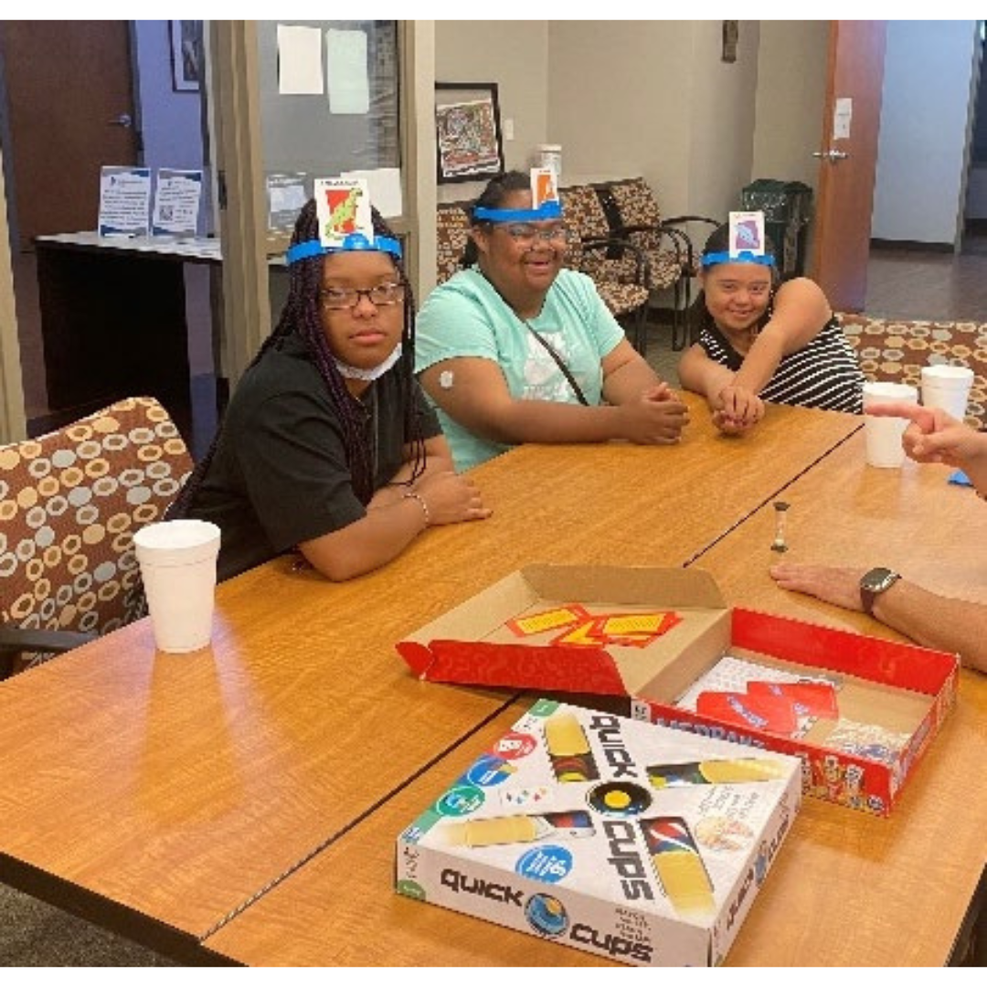 Three board game participants wear headbands with cards on them at a table with board game boxes