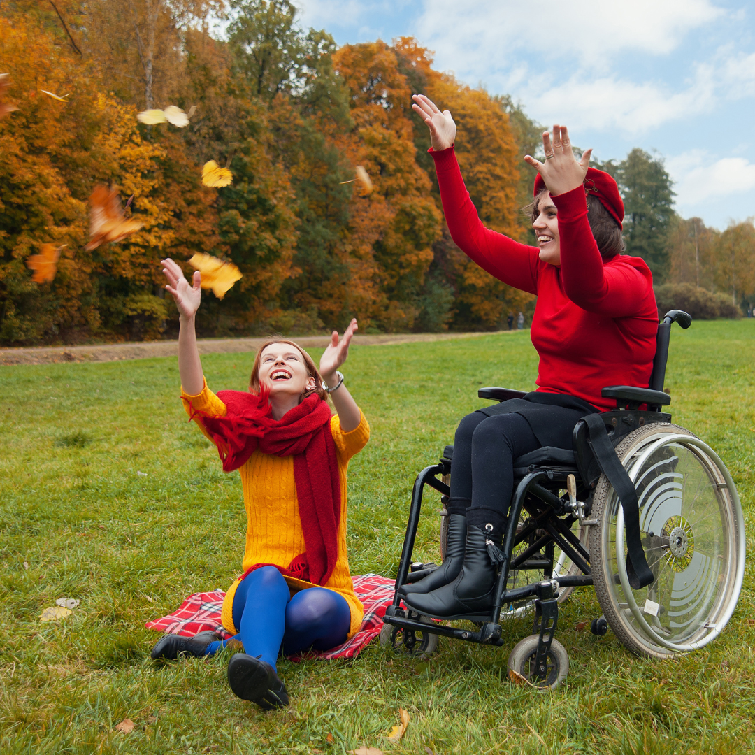 Sisters throw autumn leaves in the air. One sibling is using a wheelchair